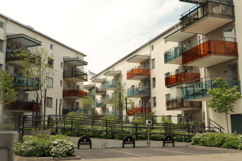 Ljusa flerbostadshus med balkongräcken i orange och grönt glas samt planteringar mellan husen.